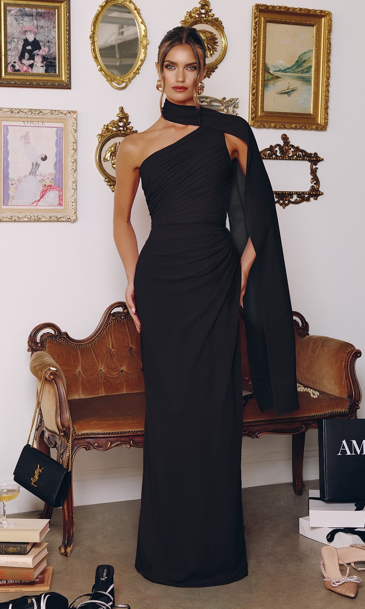 A woman wearing the Amoris T730 long black prom dress with a ruched bodice stands in a stylish room featuring vintage decor like a velvet sofa, framed art, gold mirrors, and designer shopping bags.